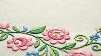 Delicate pink floral embroidery with green leaves and blue accents on cream fabric. Traditional needlework pattern for textile and craft projects.