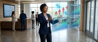 Young woman with dark hair and medium skin tone wearing a dark blazer, presenting business analytics and growth charts on a glass board in a modern office environment with colleagues in background.