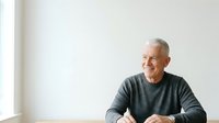 Senior man with gray hair and light skin tone, wearing dark gray long-sleeve shirt, smiling while sitting at table against white wall with natural window light for portrait and lifestyle use.