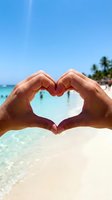 Hands forming heart shape frame tropical beach paradise with turquoise water and palm trees for vacation romance and summer love concept.