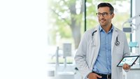 Professional male doctor with stethoscope and tablet in modern hospital corridor, smiling confidently while providing healthcare services.