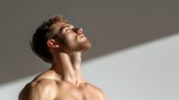 Young Caucasian male with beard tilting head back in dramatic lighting against neutral wall, showing neck and bare shoulders, expressing serenity and contemplation.