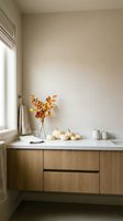 Modern minimalist kitchen counter with autumn decor featuring white pumpkins, dried branches in vase, and ceramic mugs on wooden cabinets by window.