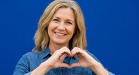 Smiling middle-aged Caucasian woman making heart shape with hands wearing denim shirt against bright blue background for wellness campaigns.