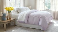 Cozy bedroom interior with lavender bedding, yellow flowers on nightstand, natural light streaming through windows creating peaceful morning atmosphere.