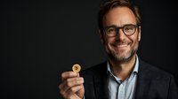 Smiling middle-aged Caucasian businessman in glasses and suit jacket holding golden bitcoin cryptocurrency coin against dark background. Financial technology concept.