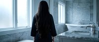 Woman with long dark hair in black robe standing by marble bathroom window overlooking luxury bathtub in modern spa-like interior for wellness relaxation.