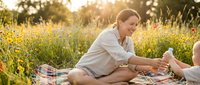 Young adult male with light skin tone and brown hair, wearing casual beige shirt, sitting on checkered blanket in sunny wildflower meadow during golden hour, engaging in outdoor leisure activity with family.