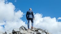 Businessman in formal suit standing on mountain peak against blue sky with clouds, symbolizing achievement, leadership and career success.