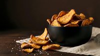 Crispy golden potato chips in black bowl on rustic wooden table with scattered salt crystals and dramatic lighting for food photography and culinary designs.