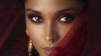 Young African woman with dramatic burgundy eye makeup, gold septum piercing and earrings against dark red background, soft focus beauty portrait highlighting flawless skin.