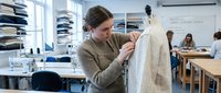 Young female fashion designer examining white garment on dress form in bright modern studio workspace with fabric shelves and sewing equipment.