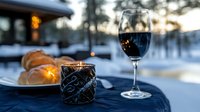 Romantic winter evening setting with red wine glass and glowing candle on outdoor table, fresh croissants in background against snowy landscape at dusk.