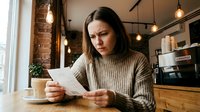 Young woman with long dark hair and olive complexion, wearing a beige ribbed sweater, reading a letter or document with concerned expression while holding a coffee cup in a modern loft kitchen with exposed brick and pendant lighting.