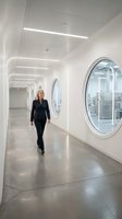 Woman in black business suit walking through modern white corridor with linear ceiling lights and circular windows, representing corporate office environment, professional workplace, and contemporary architecture design.