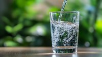 Fresh clean water pouring into clear glass on wooden table with green nature background for healthy lifestyle and hydration concepts.