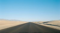 Straight asphalt road stretching through vast desert landscape with golden sand dunes and distant mountains under clear blue sky, suitable for travel and journey concepts.