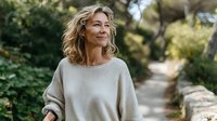 Mature woman with wavy blonde hair wearing beige sweater walking on forest path, enjoying peaceful nature moment outdoors.