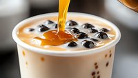 Pouring golden caramel syrup into creamy bubble milk tea with black tapioca pearls, close-up view showing rich texture and flowing liquid motion.