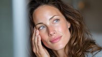 Young woman with wavy brown hair and natural makeup touching her face gently, looking thoughtfully with soft expression in natural light setting.