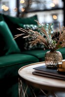Cozy home interior with emerald velvet sofa, round wooden coffee table and decorative dried flowers in metal vase. Soft bokeh lights create warm atmosphere.
