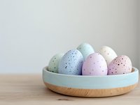 Speckled pastel Easter eggs in blue, pink, and mint colors arranged in modern wooden bowl with turquoise rim on wooden surface against neutral wall background.
