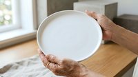 Senior adult with medium skin tone holding empty white ceramic plate in bright kitchen setting, displaying dishware for dining or food presentation purposes.