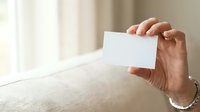 Adult male hand holding blank white business card against neutral interior background with natural window light.