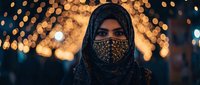 Young Middle Eastern woman wearing ornate face mask and hijab with golden bokeh lights background creating warm atmospheric portrait for cultural diversity concepts.