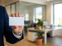 Businessman in dark suit holding blank white business card in modern office environment with blurred background for corporate branding and contact information.