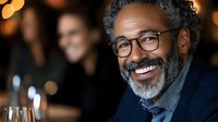 Mature African American male professional wearing eyeglasses and dark blazer smiling at corporate event or business gathering with blurred colleagues in background.