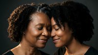 African American mother and adult daughter sharing intimate tender moment with foreheads touching in warm emotional embrace against dark background.