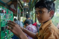 Young South Asian boy working on electronic circuit board in workshop environment with other technicians in background.