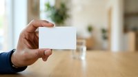 Male professional in business attire holding blank white card at wooden table in modern office setting, suitable for corporate branding, business card mockups, and professional presentations.