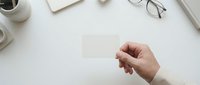 Hand holding blank white business card mockup on clean desk workspace with coffee cup, notebook and glasses for branding presentation.