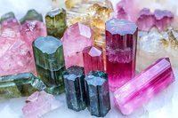 Raw tourmaline crystals in various colors from pink to green and blue, showing natural prismatic formations and mineral structure on white background.