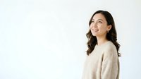 Young woman with long dark wavy hair and East Asian ethnicity wearing a beige cardigan, smiling while looking upward against white background for portrait and lifestyle use.