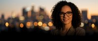 Young woman with curly dark hair and glasses smiling at camera against blurred city skyline at dusk, suitable for corporate portraits and professional headshots.