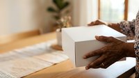 Person with dark skin tone holding white cardboard box on wooden table indoors, suitable for e-commerce, shipping, delivery, unboxing, and product packaging concepts.