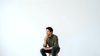 Young man with dark curly hair and olive skin tone wearing olive green shirt, crouching against white background, contemplative pose for portrait and lifestyle design use.