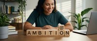 Woman with dark hair and medium skin tone, wearing a teal t-shirt, smiling while arranging wooden blocks spelling ambition at desk with laptop and plants.
