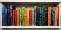 Colorful vintage hardcover books arranged on white wooden shelf, spines showing rainbow spectrum arrangement from blue to orange, creating organized library display.