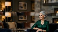 Senior woman with gray hair and light skin tone, wearing elegant green blouse, sitting on dark sofa in luxury living room with warm lighting, fireplace, and modern decor for lifestyle and home design.