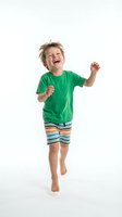 Energetic young boy jumping with joy in green shirt and striped shorts on white background. Happy child celebrating success with arms raised and big smile.