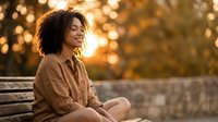 Young African American woman with curly hair sitting on wooden bench during golden hour sunset, smiling peacefully in warm autumn light for lifestyle content.