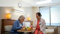 Healthcare worker assists elderly woman with meal in bright hospital room. Caregiver helps senior patient eat nutritious food during recovery treatment.