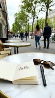 Open notebook with hello written inside, sunglasses and pen on marble cafe table, Parisian street with pedestrians and trees in background.