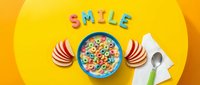Colorful cereal loops in a bowl with milk, apple slices, spoon, and the word SMILE made of letter candies on a bright yellow background.