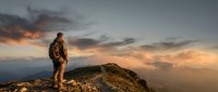 Male hiker with backpack standing on mountain summit at sunset, overlooking vast landscape with dramatic cloudy sky and golden light for travel and adventure concepts.
