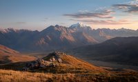 Majestic mountain peaks at golden sunset with dramatic light illuminating rugged terrain and alpine meadows, creating atmospheric landscape in wilderness setting.